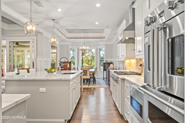 kitchen featuring hanging light fixtures, a kitchen island with sink, a sink, wall chimney range hood, and white cabinets