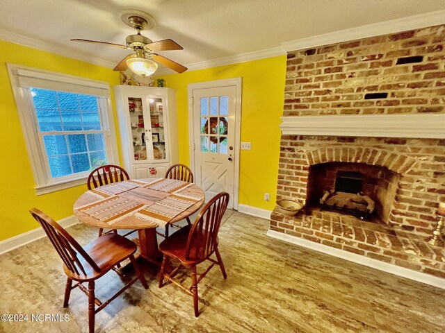 dining room featuring wood finished floors, a ceiling fan, baseboards, ornamental molding, and a brick fireplace