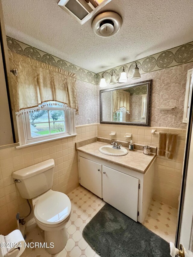 bathroom featuring wallpapered walls, toilet, a wainscoted wall, a textured ceiling, and tile walls