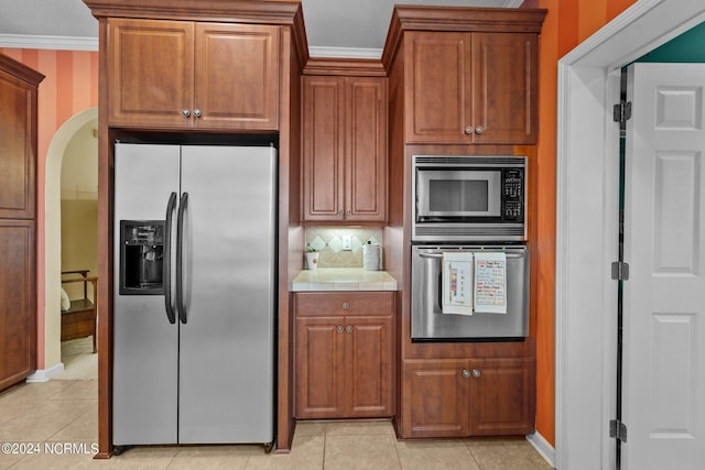 kitchen with arched walkways, tile countertops, appliances with stainless steel finishes, brown cabinets, and ornamental molding