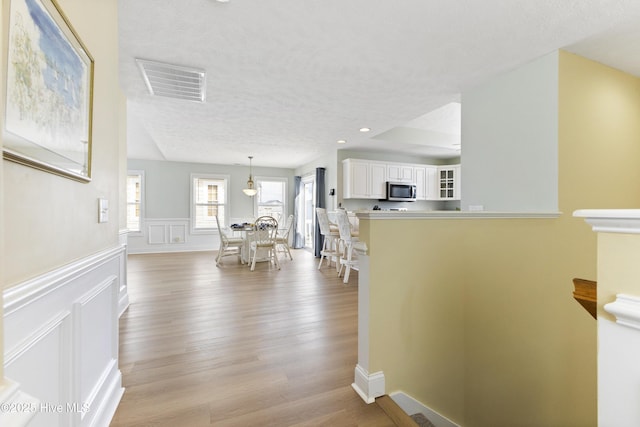 hallway featuring visible vents, wainscoting, a textured ceiling, light wood-type flooring, and a decorative wall