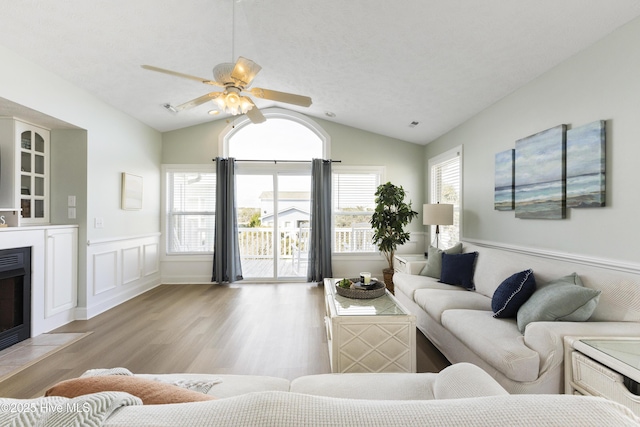 living room featuring ceiling fan, a wainscoted wall, a fireplace with flush hearth, vaulted ceiling, and light wood finished floors