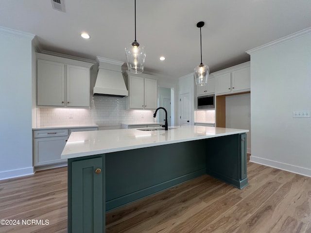 kitchen featuring custom exhaust hood, a center island with sink, light wood-type flooring, sink, and decorative light fixtures