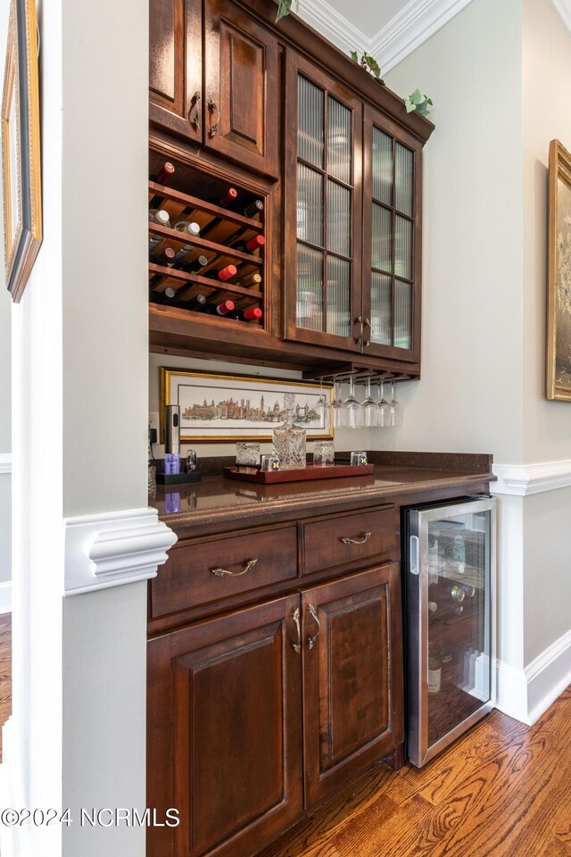 bar featuring ornamental molding, dark brown cabinets, beverage cooler, and hardwood / wood-style flooring