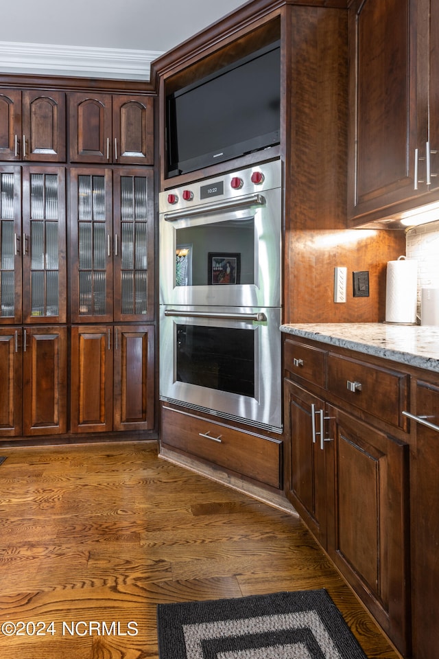 kitchen featuring stainless steel double oven, light stone counters, dark hardwood / wood-style floors, dark brown cabinetry, and ornamental molding