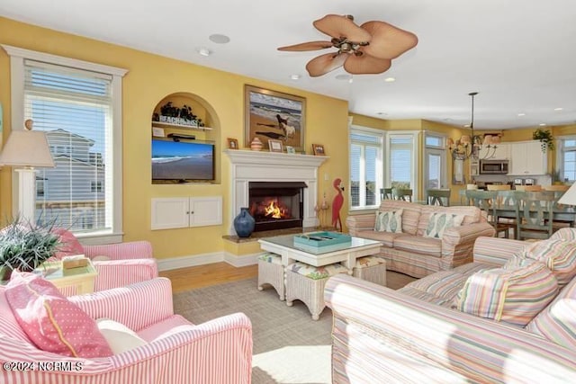 living room featuring baseboards, light wood-style flooring, a lit fireplace, ceiling fan with notable chandelier, and recessed lighting