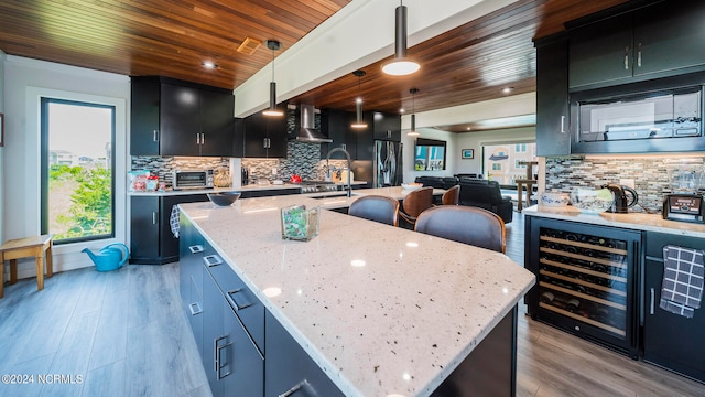 kitchen featuring beverage cooler, light hardwood / wood-style flooring, decorative light fixtures, a kitchen island with sink, and wall chimney range hood