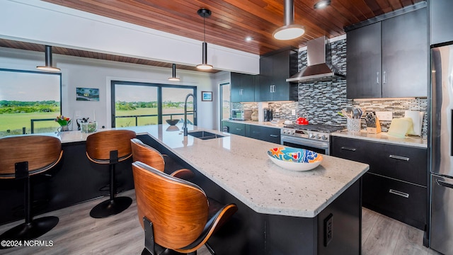 kitchen featuring pendant lighting, a center island with sink, wall chimney range hood, a kitchen breakfast bar, and appliances with stainless steel finishes