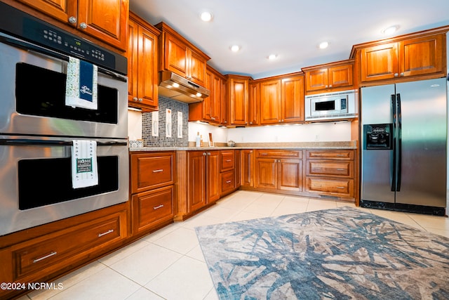 kitchen with light tile patterned floors, appliances with stainless steel finishes, brown cabinets, light stone counters, and under cabinet range hood