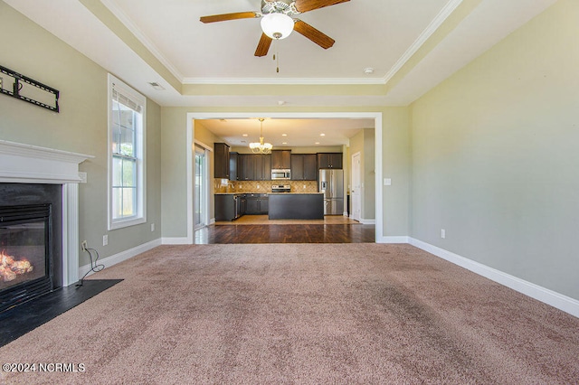 unfurnished living room featuring crown molding, dark colored carpet, ceiling fan with notable chandelier, and a raised ceiling