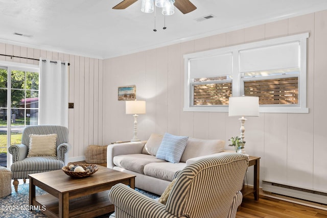 living room with a baseboard heating unit, wood-type flooring, wood walls, crown molding, and ceiling fan