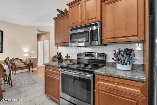 kitchen featuring dark countertops, stainless steel appliances, light tile patterned flooring, brown cabinetry, and decorative backsplash