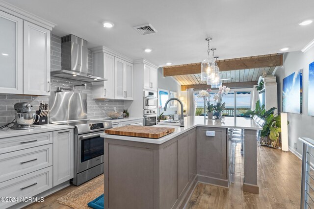 kitchen featuring wall chimney exhaust hood, stainless steel appliances, an island with sink, and white cabinets