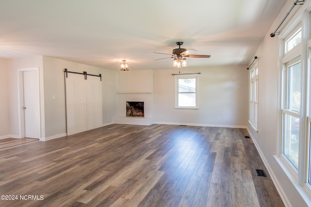 unfurnished living room with a brick fireplace, ceiling fan, a barn door, and dark hardwood / wood-style floors