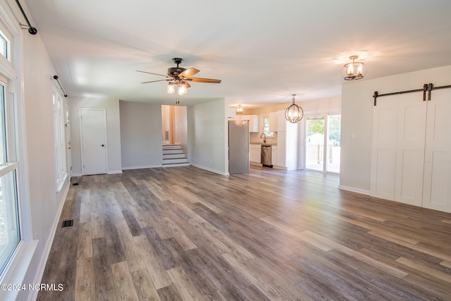 unfurnished living room with a barn door, ceiling fan with notable chandelier, wood-type flooring, and sink