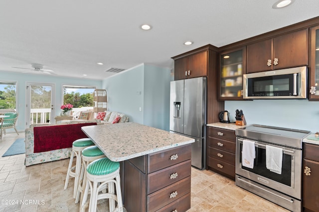 kitchen with light stone countertops, dark brown cabinets, appliances with stainless steel finishes, a center island, and ceiling fan