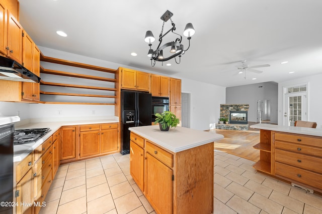 kitchen featuring a center island, a stone fireplace, decorative light fixtures, black appliances, and ceiling fan with notable chandelier