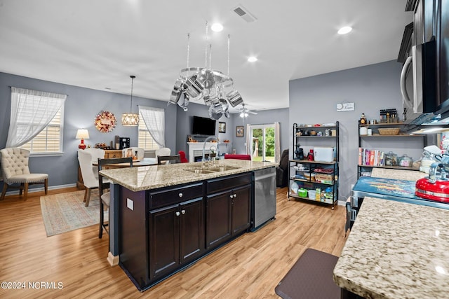 kitchen featuring stainless steel appliances, light stone counters, a center island with sink, decorative light fixtures, and light wood-type flooring