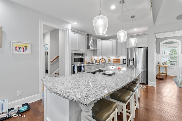 kitchen with wall chimney range hood, hanging light fixtures, stainless steel appliances, a center island with sink, and sink