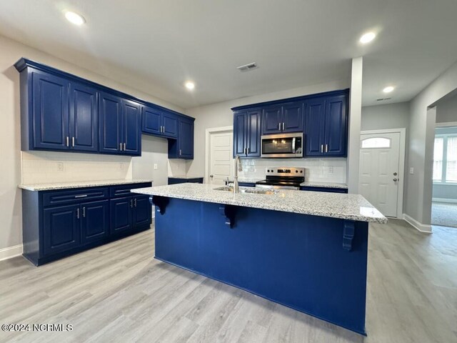 kitchen with appliances with stainless steel finishes, blue cabinets, a breakfast bar area, light wood-type flooring, and a kitchen island with sink