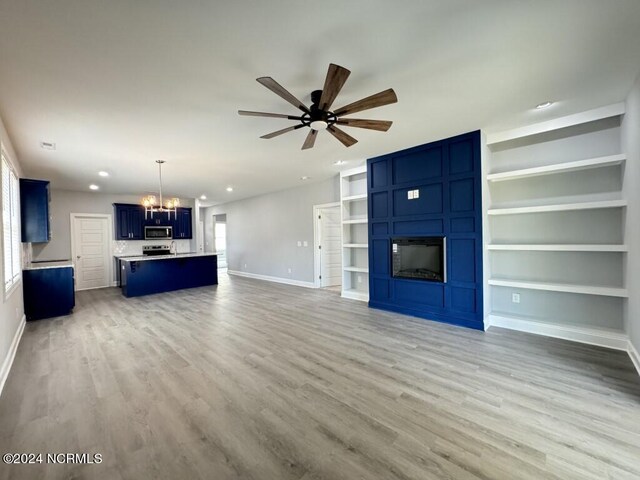 unfurnished living room with built in shelves, ceiling fan with notable chandelier, and light hardwood / wood-style floors