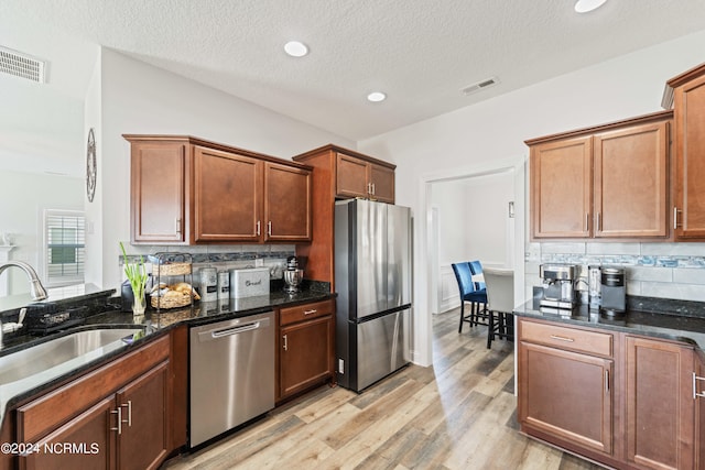 kitchen with light wood-type flooring, a textured ceiling, stainless steel appliances, backsplash, and dark stone countertops