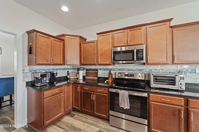 kitchen featuring dark stone countertops, backsplash, stainless steel appliances, a textured ceiling, and light hardwood / wood-style flooring