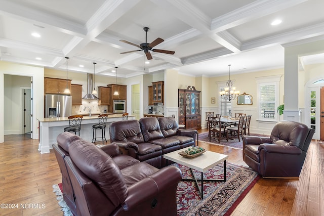 living room with coffered ceiling, ceiling fan with notable chandelier, sink, ornamental molding, and beam ceiling