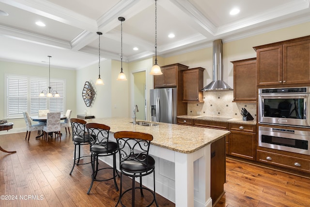 kitchen with a kitchen island with sink, wall chimney range hood, sink, hanging light fixtures, and stainless steel appliances