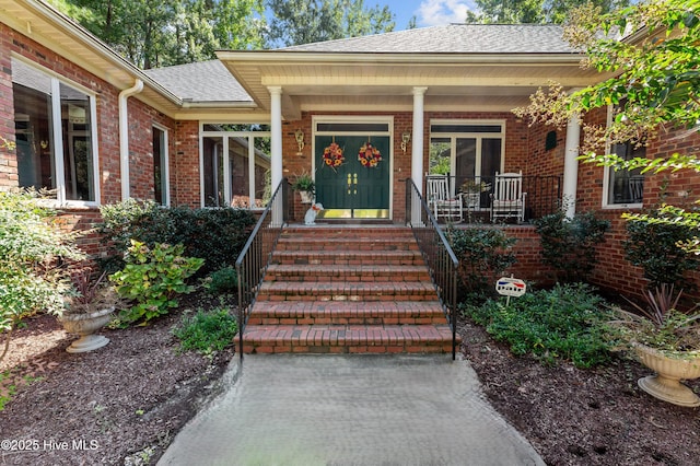 entrance to property featuring covered porch, brick siding, and roof with shingles