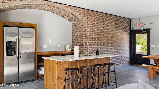 bar with light stone counters, brick wall, sink, and stainless steel refrigerator with ice dispenser