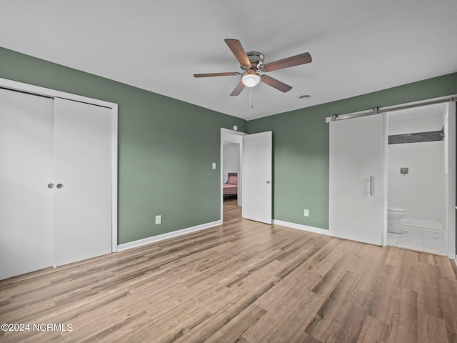 unfurnished bedroom featuring ceiling fan, light hardwood / wood-style flooring, a closet, a barn door, and ensuite bathroom