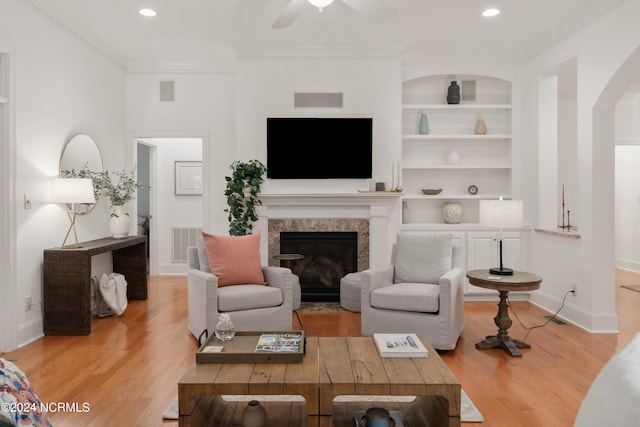 living room with a high end fireplace, crown molding, ceiling fan, built in features, and light hardwood / wood-style floors