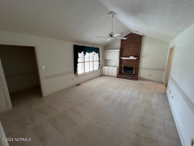 unfurnished living room with vaulted ceiling, a brick fireplace, a textured ceiling, ceiling fan, and light colored carpet