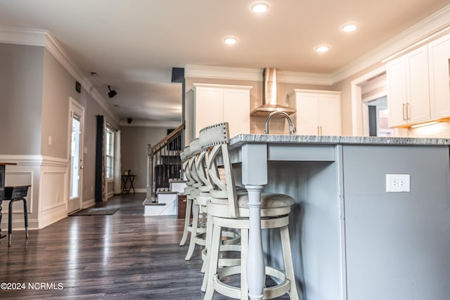 kitchen featuring white cabinetry, a kitchen bar, wall chimney range hood, dark hardwood / wood-style flooring, and ornamental molding