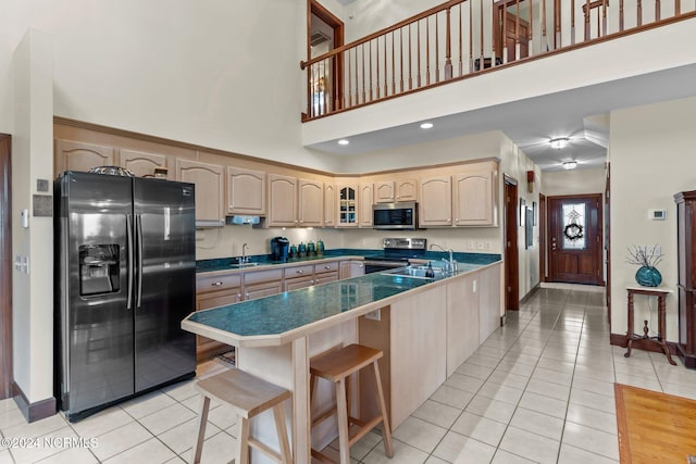 kitchen featuring sink, a towering ceiling, appliances with stainless steel finishes, light brown cabinetry, and a kitchen bar