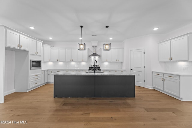 kitchen featuring stainless steel microwave, a kitchen island with sink, light hardwood / wood-style flooring, white cabinets, and hanging light fixtures