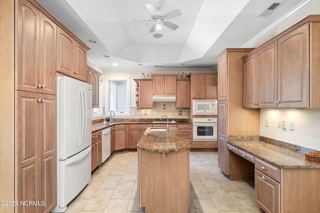 kitchen with white appliances, a kitchen island with sink, sink, a tray ceiling, and light stone counters