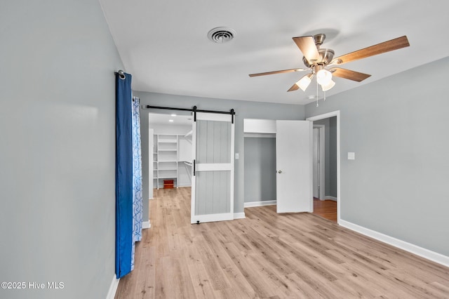 unfurnished bedroom with ceiling fan, a barn door, a walk in closet, a closet, and light wood-type flooring