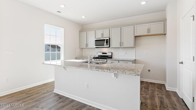 kitchen featuring appliances with stainless steel finishes, light stone counters, dark hardwood / wood-style flooring, gray cabinets, and a center island with sink