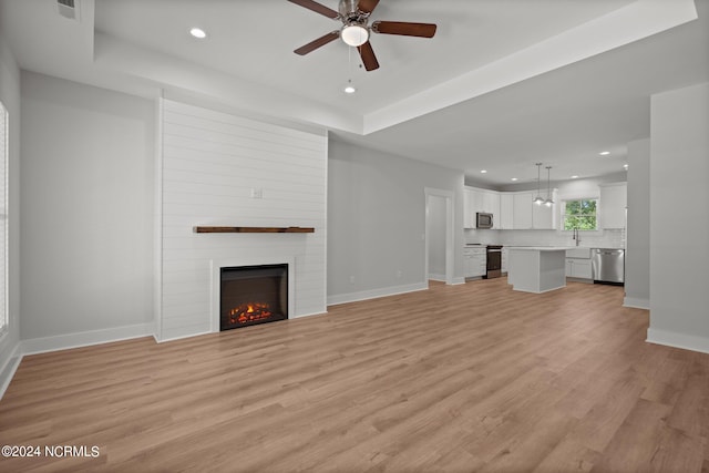 unfurnished living room featuring ceiling fan, a large fireplace, a raised ceiling, and light hardwood / wood-style floors