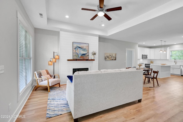 living room with a raised ceiling, ceiling fan, a fireplace, and light hardwood / wood-style flooring