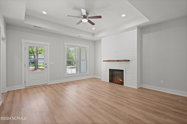 unfurnished living room featuring ceiling fan, a large fireplace, a tray ceiling, and light hardwood / wood-style floors