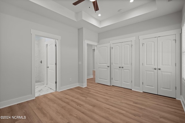 unfurnished bedroom featuring ensuite bathroom, light wood-type flooring, multiple closets, a tray ceiling, and ceiling fan