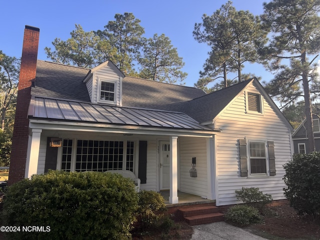 cape cod house with a standing seam roof, a porch, a chimney, metal roof, and a shingled roof