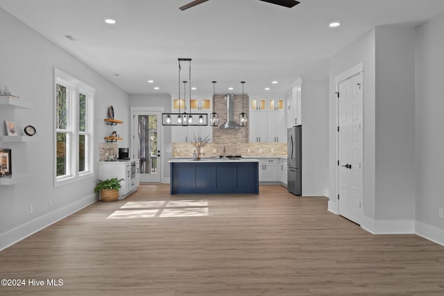 kitchen featuring hanging light fixtures, wall chimney exhaust hood, tasteful backsplash, white cabinetry, and stainless steel refrigerator