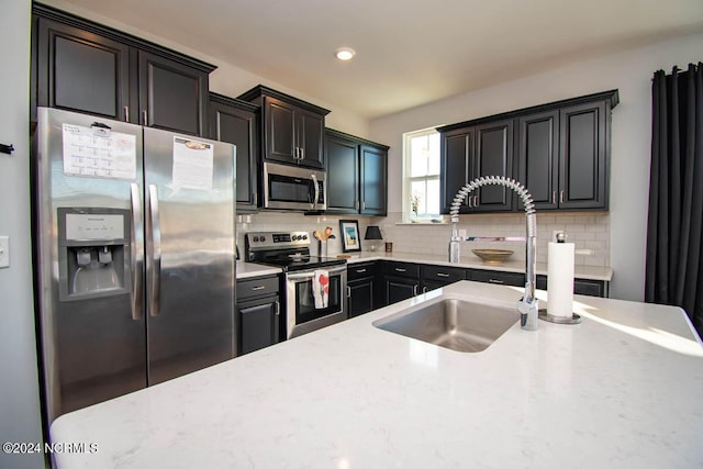 kitchen with light stone countertops, a sink, dark cabinetry, stainless steel appliances, and decorative backsplash