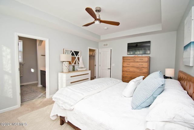 bedroom featuring light carpet, baseboards, ensuite bathroom, a raised ceiling, and a ceiling fan