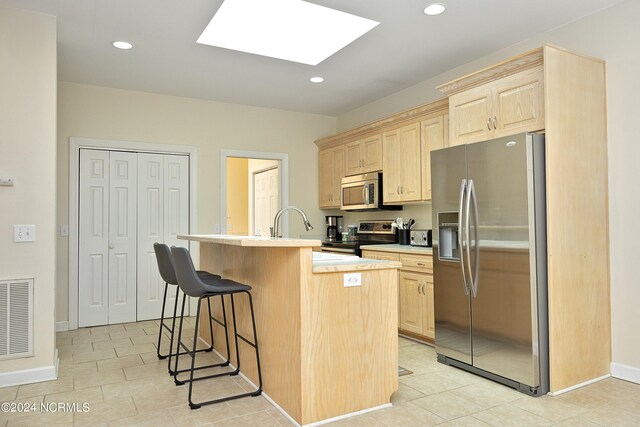 kitchen featuring a skylight, light brown cabinetry, an island with sink, appliances with stainless steel finishes, and a kitchen bar