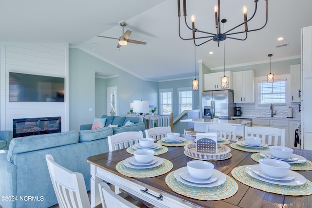 dining space featuring sink, ceiling fan with notable chandelier, crown molding, vaulted ceiling, and a large fireplace